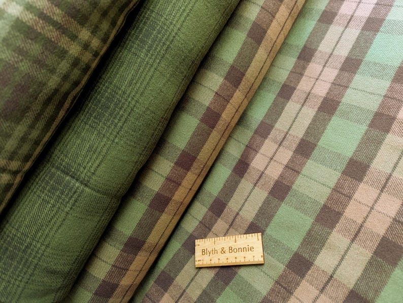 May include: Close-up of several rolls of plaid fabric in shades of green, brown, and tan. The fabric has a classic plaid pattern with intersecting lines. A wooden ruler with the text "Blyth & Bonnie" is visible.