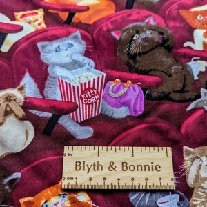 May include: Fabric featuring a pattern of cats sitting in red theater seats, with a red and white striped popcorn bucket labeled Kitty Corn, a purple purse, and a wooden ruler measuring 7 inches long placed across the center of the design.