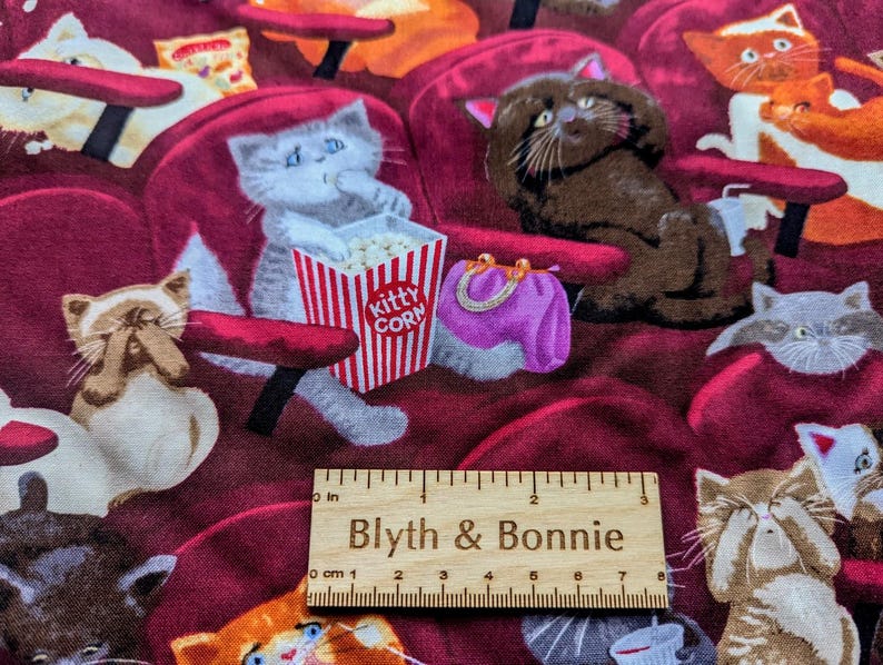 May include: Fabric featuring a pattern of cats sitting in red theater seats, with a red and white striped popcorn bucket labeled Kitty Corn, a purple purse, and a wooden ruler measuring 7 inches long placed across the center of the design.