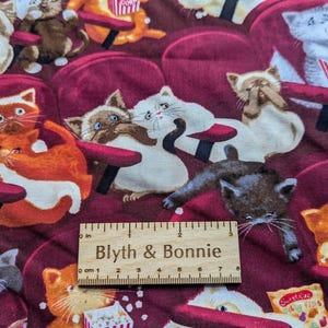 May include: Fabric featuring a pattern of various cats sitting in dark red theater seats, some eating popcorn from striped containers. A wooden ruler measuring 6 inches is placed horizontally across the center of the fabric print.