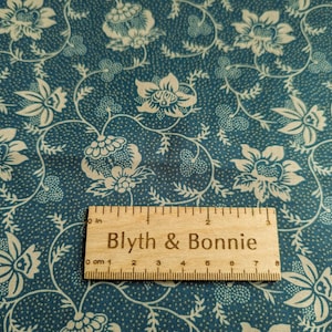 Puede incluir: Primer plano de una tela color turquesa con un estampado floral crema. El dise&ntilde;o presenta flores y hojas estilizadas sobre un fondo punteado. Una regla de madera con el texto "Blyth & Bonnie" mide hasta 7 cm.