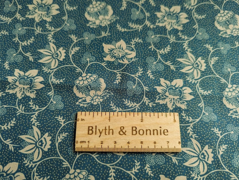 Puede incluir: Primer plano de una tela color turquesa con un estampado floral crema. El dise&ntilde;o presenta flores y hojas estilizadas sobre un fondo punteado. Una regla de madera con el texto "Blyth & Bonnie" mide hasta 7 cm.