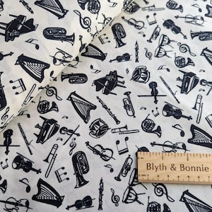 May include: A white fabric with a black musical instrument pattern. The pattern includes a variety of instruments such as a trumpet, a harp, a tuba, a violin, a drum, and a piano. The fabric also includes musical notes and a ruler with the text "Blyth & Bonnie".