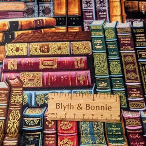 May include: A fabric print depicting a library of antique books in shades of burgundy, gold, and teal. The books are adorned with gold detailing and arranged in a stacked pattern. A wooden ruler with the text "Blyth & Bonnie" is visible in the foreground.