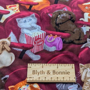May include: A fabric print featuring various cats sitting in red theater seats, including a gray cat eating from a red and white striped box labeled Kitty Corn and a purple purse. A wooden ruler shows 3 inches of length at the bottom center.