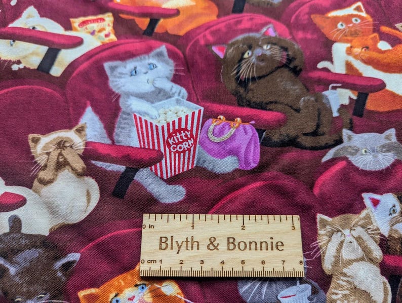 May include: A fabric print featuring various cats sitting in red theater seats, including a gray cat eating from a red and white striped box labeled Kitty Corn and a purple purse. A wooden ruler shows 3 inches of length at the bottom center.