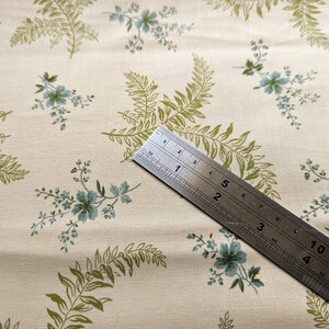 Pode incluir: Um close-up de um tecido cor de creme com um padr&atilde;o repetitivo de folhas de samambaia verdes e flores azuis. Uma r&eacute;gua de metal &eacute; colocada diagonalmente sobre o tecido, com os n&uacute;meros de 1 a 5 vis&iacute;veis.