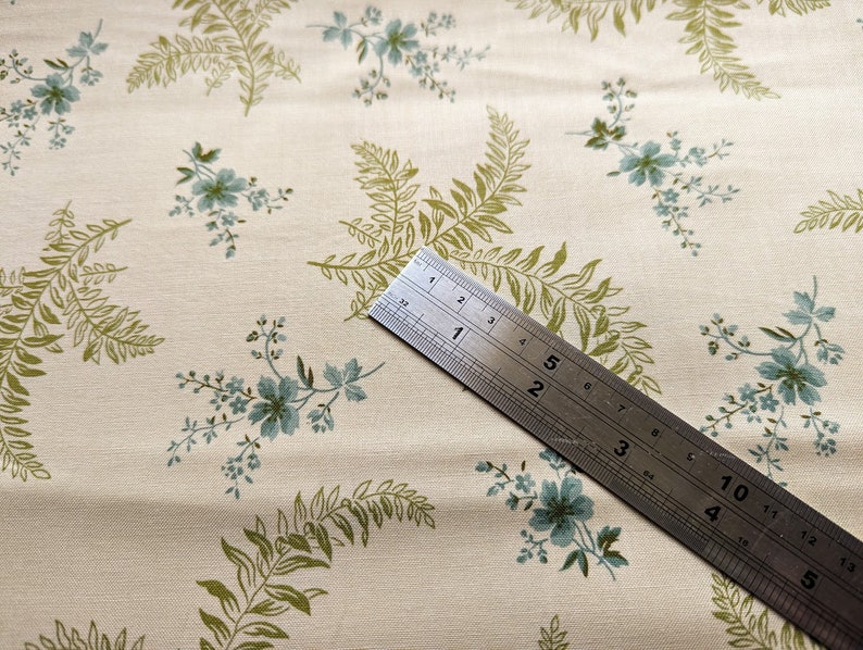 Pode incluir: Um close-up de um tecido cor de creme com um padr&atilde;o repetitivo de folhas de samambaia verdes e flores azuis. Uma r&eacute;gua de metal &eacute; colocada diagonalmente sobre o tecido, com os n&uacute;meros de 1 a 5 vis&iacute;veis.