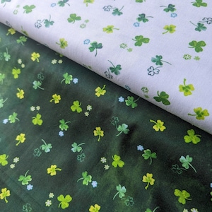 Op de afbeelding: Twee stofstalen met een groen klaverbladpatroon. De bovenste staal is wit met groene klaverbladeren. De onderste staal is donkergroen met lichtgroene klaverbladeren.