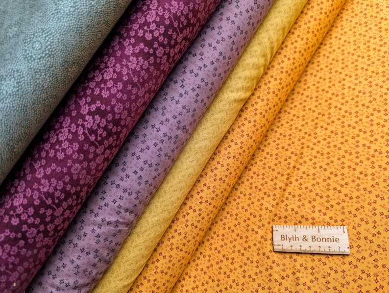 Puede incluir: Rollos de tela en varios colores, incluyendo verde azulado, morado oscuro, lavanda, amarillo y naranja. Las telas presentan patrones florales y geom&eacute;tricos. Se ve una regla de madera con el texto "Blyth & Bonnie".