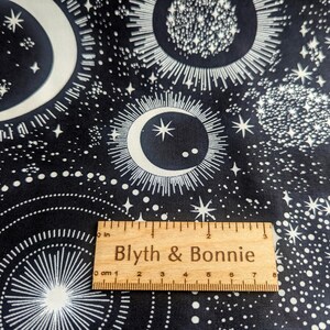 May include: Black fabric with a white celestial pattern featuring moons, stars, and suns. A wooden ruler with the text "Blyth & Bonnie" is placed on the fabric. The ruler measures up to 8 inches.