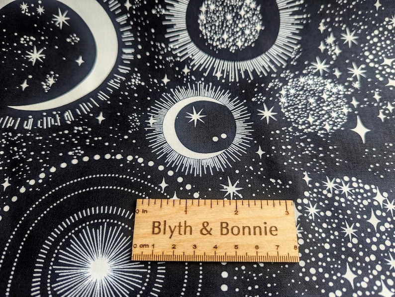 May include: Black fabric with a white celestial pattern featuring moons, stars, and suns. A wooden ruler with the text "Blyth & Bonnie" is placed on the fabric. The ruler measures up to 8 inches.