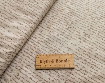 Pode incluir: Um tecido bege com um padrão de escrita marrom. O tecido tem uma régua em cima com as palavras "Blyth & Bonnie" impressas nela. A régua tem marcas de 1 a 9.