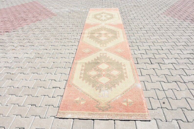 Puede incluir: Una alfombra larga y rectangular con un patr&oacute;n geom&eacute;trico en tonos melocot&oacute;n, beige y marr&oacute;n. La alfombra tiene un aspecto desgastado y vintage.