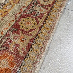 Puede incluir: Primer plano de una alfombra beige y naranja con estampado y borde rojo y negro. La alfombra est&aacute; sobre un suelo de madera claro.