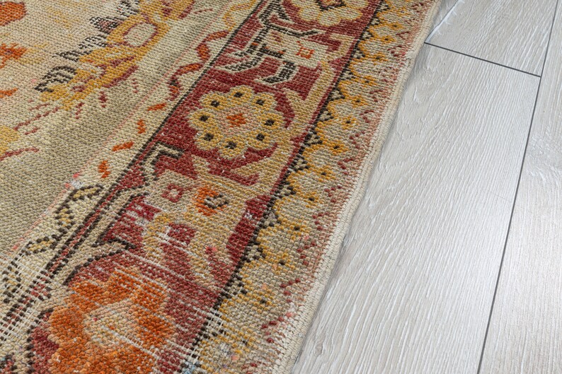 Puede incluir: Primer plano de una alfombra beige y naranja con estampado y borde rojo y negro. La alfombra est&aacute; sobre un suelo de madera claro.