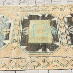 May include: A vintage rug with a beige background and intricate patterns in shades of brown, blue, and green. The rug features a central design with a square shape and a border of geometric motifs.