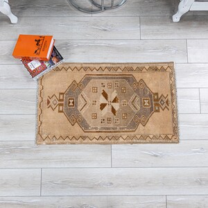 May include: A rectangular, beige and brown patterned rug with a central geometric design. The rug features a central medallion with a cross-like pattern and is surrounded by intricate borders. Two books are on top of the rug.