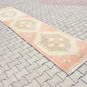 Puede incluir: Una alfombra larga y rectangular con fondo crema y un patr&oacute;n geom&eacute;trico en tonos melocot&oacute;n, marr&oacute;n y beige. La alfombra est&aacute; colocada sobre una superficie de ladrillo gris.