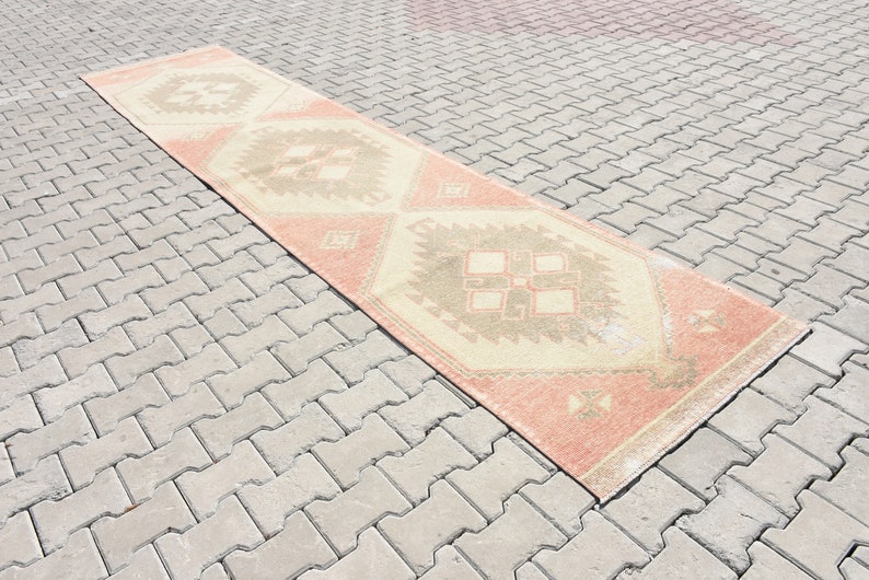 Puede incluir: Una alfombra larga y rectangular con fondo crema y un patr&oacute;n geom&eacute;trico en tonos melocot&oacute;n, marr&oacute;n y beige. La alfombra est&aacute; colocada sobre una superficie de ladrillo gris.