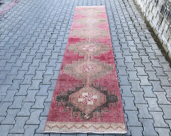 Puede incluir: Una alfombra larga y estrecha con un diseño turco tradicional. La alfombra es principalmente roja con acentos beige, verde y negro. La alfombra tiene un patrón geométrico con una forma de diamante central.
