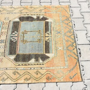 May include: A vintage rug with a geometric pattern in shades of beige, brown, blue, and pink. The rug has a fringe border and is laid out on a gray concrete surface.