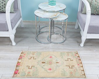 May include: A beige rug with a light green and pink floral pattern. The rug is placed in front of a set of three nesting tables with a white marble top and silver metal legs. The tables are decorated with a teal and white geometric patterned vase.