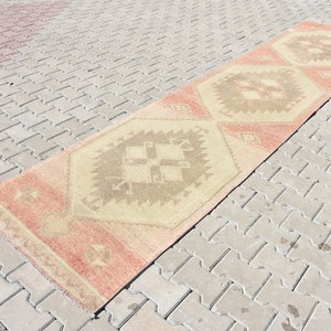 Puede incluir: Una alfombra larga y rectangular con fondo beige y un patr&oacute;n geom&eacute;trico en tonos marrones, rosas y crema. La alfombra est&aacute; colocada sobre una superficie de ladrillo gris.