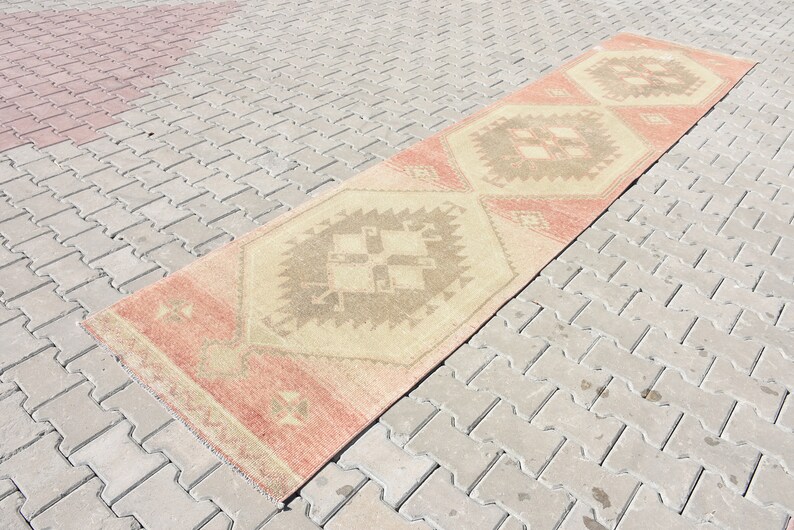 Puede incluir: Una alfombra larga y rectangular con fondo beige y un patr&oacute;n geom&eacute;trico en tonos marrones, rosas y crema. La alfombra est&aacute; colocada sobre una superficie de ladrillo gris.
