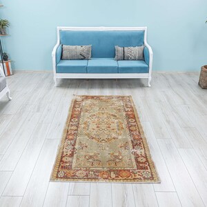 Puede incluir: Una alfombra beige claro con un patr&oacute;n floral en tonos marrones, naranjas y rojos. La alfombra est&aacute; colocada sobre un suelo de madera clara en una habitaci&oacute;n con un sof&aacute; blanco y una planta en maceta.