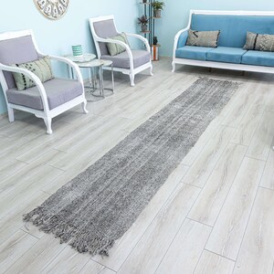 May include: A gray and white runner rug with fringe edges. The rug is laid out on a light wood floor in a living room setting with white furniture.