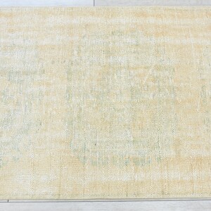 May include: A light beige rug with a faded green pattern in the center. The rug has a distressed look and is made of a soft, woven material.