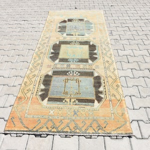 May include: A vintage Turkish runner rug with a geometric pattern in shades of beige, blue, and brown. The rug has a fringe border and is laid out on a gray concrete surface.