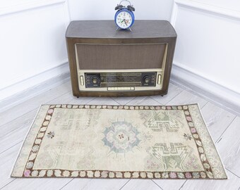 May include: A vintage-style rug with a light beige background and a brown border. The rug has a geometric pattern in shades of green and pink.