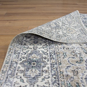 May include: A close-up of a patterned rug with a cream background and blue, gray, and brown accents. The rug is folded over, revealing the underside of the fabric.
