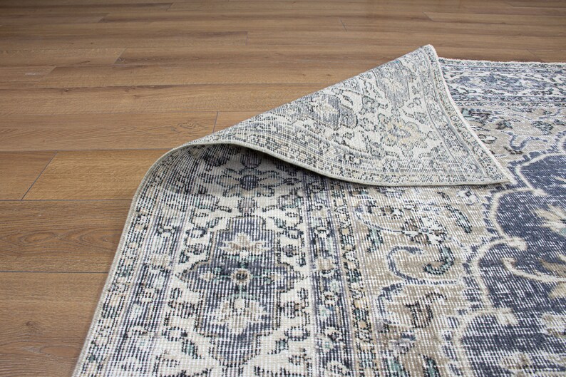 May include: A close-up of a patterned rug with a cream background and blue, gray, and brown accents. The rug is folded over, revealing the underside of the fabric.