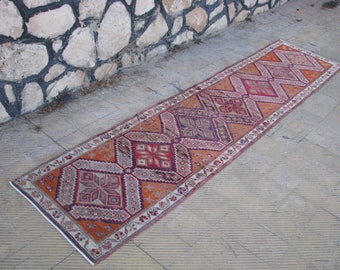 Könnte beinhalten: Ein langer, schmaler, orangefarbener und roter Teppich mit geometrischem Muster. Der Teppich hat einen weißen Rand und liegt auf einem Betonboden.