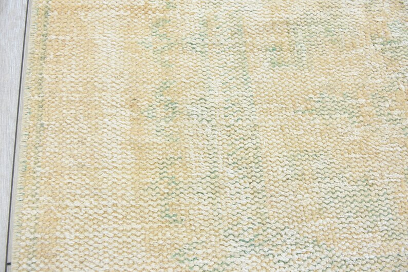 May include: Close-up of a cream-colored woven rug with a subtle green pattern. The rug has a textured surface and a slightly distressed look.