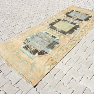 May include: A vintage Turkish rug with a light beige background and a geometric pattern in shades of blue, brown, and white. The rug has a fringed edge and is laid out on a gray brick surface.