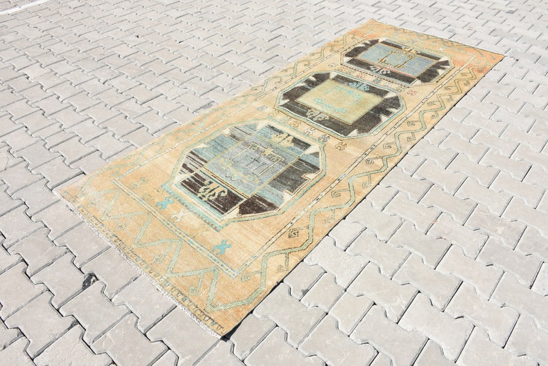 May include: A vintage Turkish rug with a light beige background and a geometric pattern in shades of blue, brown, and white. The rug has a fringed edge and is laid out on a gray brick surface.
