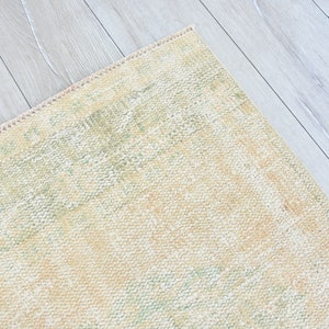 May include: A close-up of a light beige rug with a subtle green and cream pattern. The rug has a fringed edge and is lying on a light wood floor.