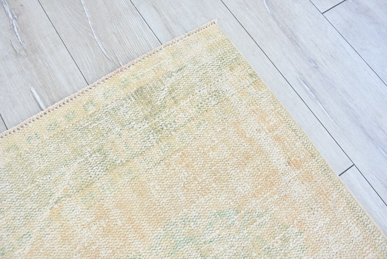 May include: A close-up of a light beige rug with a subtle green and cream pattern. The rug has a fringed edge and is lying on a light wood floor.