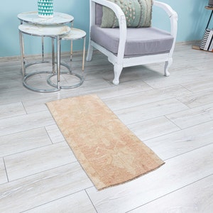 May include: A light beige runner rug with a faded floral pattern. The rug is laid out on a light wood floor in a room with a white chair and a set of nesting tables.