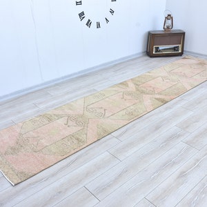 Puede incluir: Una alfombra de pasillo larga y estrecha con un fondo rosa claro y un patrón geométrico en color beige oscuro. La alfombra está colocada sobre un suelo de madera claro.