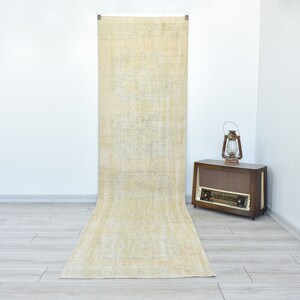 May include: A long, off-white runner rug with a subtle pattern. The rug is hanging on a white wall in a room with light wood floors.