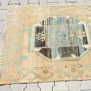 May include: A vintage rug with a beige background and a central design featuring a blue, brown, and white geometric pattern. The rug has a border with a repeating pattern of blue and white.