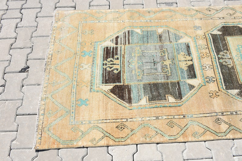 May include: A vintage rug with a beige background and a central design featuring a blue, brown, and white geometric pattern. The rug has a border with a repeating pattern of blue and white.