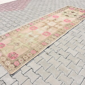 Puede incluir: Una alfombra larga y rectangular con un patrón geométrico descolorido en rosa, marrón y verde. La alfombra está colocada sobre una superficie de ladrillo gris.