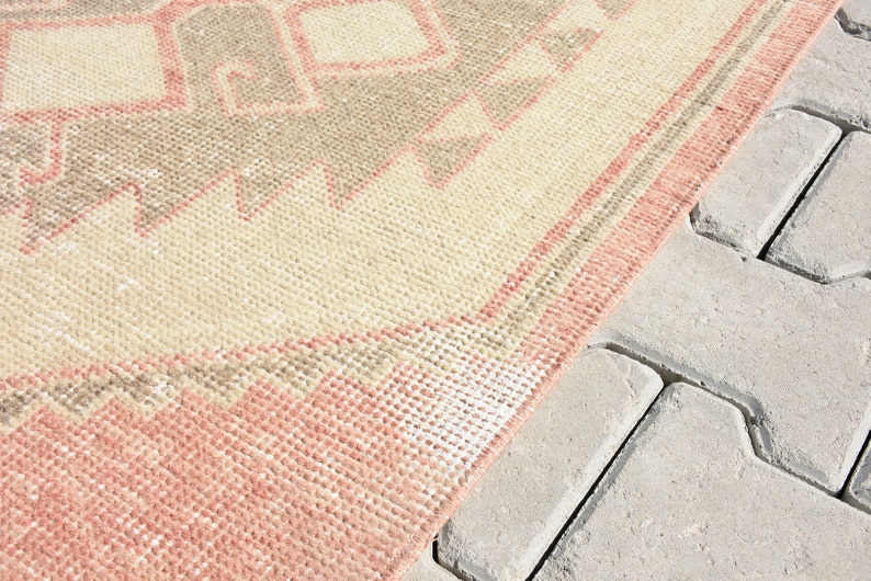 Puede incluir: Una alfombra de color rosa y beige con un dise&ntilde;o geom&eacute;trico. La alfombra est&aacute; sobre una superficie de hormig&oacute;n gris.