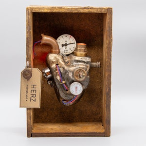 Könnte beinhalten: Eine Steampunk-Metall-Herzskulptur mit Zahnrädern, Rohren und Messgeräten, ausgestellt in einer Holzkiste mit einem Etikett, auf dem "HERZ *versteiner*t*" steht.